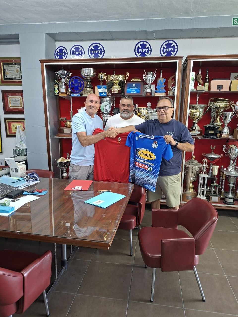 Representantes del CD Mensajero y la UD Tarajalejo se dan la mano ante una vitrina de trofeos